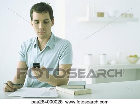 Young man at table with paper and books, looking down at cell phone View Large Photo Image Stock Photo - Young man at table with paper and books, looking down at cell phone. Fotosearch