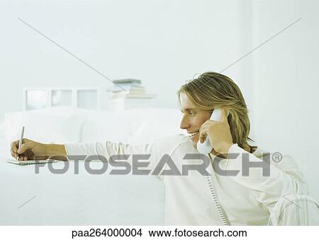 Young man holding landline phone to ear, taking notes on notepad View Large Photo Image Picture - Young man holding landline phone to ear, taking notes on notepad. Fotosearch