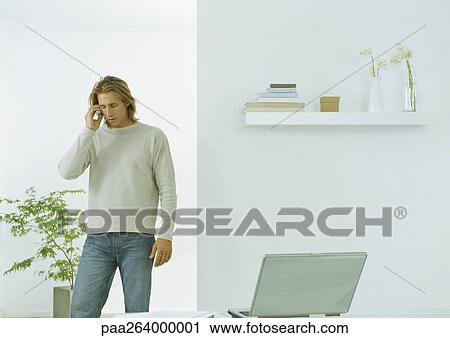 Young man in casual clothes standing with cell phone to ear, indoors View Large Photo Image Stock Image - Young man in casual clothes standing with cell phone to ear, indoors. Fotosearch