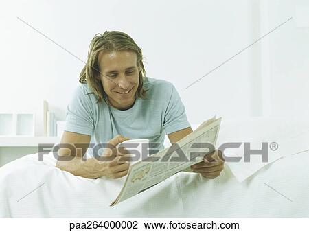 Young man lying on stomach with mug in one hand and newspaper in other View Large Photo Image Stock Image - Young man lying on stomach with mug in one hand and newspaper in other. Fotosearch