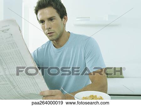 Young man sitting at table with bowl of cereal holding financial page of newspaper View Large Photo Image Stock Photo - Young man sitting at table with bowl of cereal holding financial page of newspaper. Fotosearch