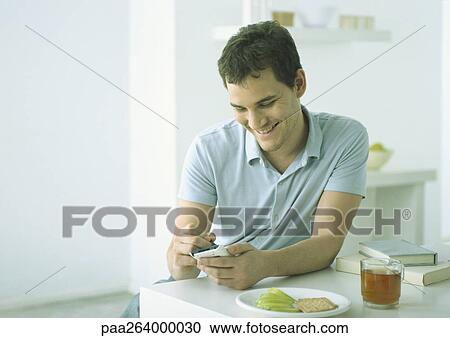 Young man sitting at table with food, working on electronic organizer View Large Photo Image Stock Image - Young man sitting at table with food, working on electronic organizer. Fotosearch