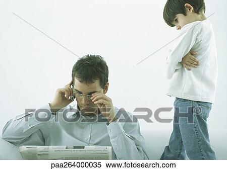 Stock Photography - Young man sitting, reading newspaper, talking on cell phone, boy looking down at him with arms crossed. Fotosearch