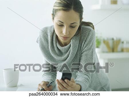 Young woman leaning on table, looking down at cell phone View Large Photo Image Picture - Young woman leaning on table, looking down at cell phone. Fotosearch