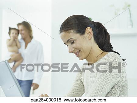 Stock Photo - Young woman looking down at laptop, young man holding little girl in background. Fotosearch