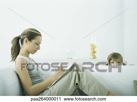 Stock Image - Young woman sitting on sofa writing on notepad, girl peeking over edge of sofa. Fotosearch