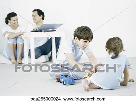 Boy and girl playing on floor, parents using laptop in background View Large Photo Image Picture - Boy and girl playing on floor, parents using laptop in background . Fotosearch