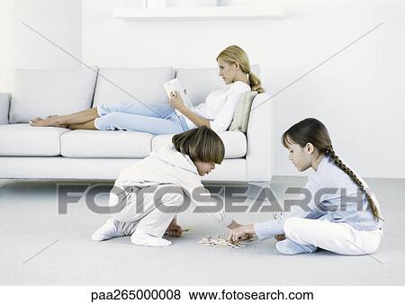 Stock Photo - Boy and girl sitting on floor playing, woman sitting on sofa reading in background. Fotosearch
