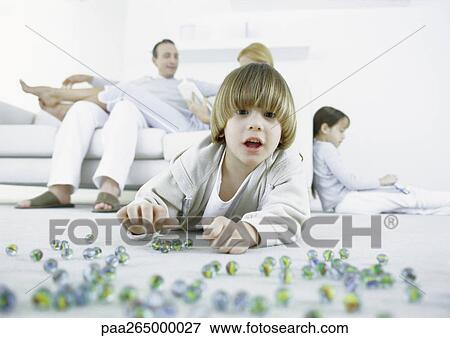 Boy lying on floor playing marbles, parents and sister sitting in background View Large Photo Image Stock Photo - Boy lying on floor playing marbles, parents and sister sitting in background. Fotosearch