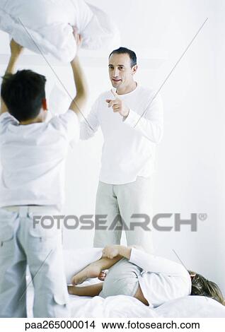 Picture - Boy standing on bed holding up pillow, rear view, second boy lying on bed, man pointing at first boy. Fotosearch
