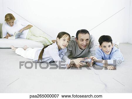 Stock Photo - Man and boy and girl lying on floor playing video game, woman reading in background. Fotosearch