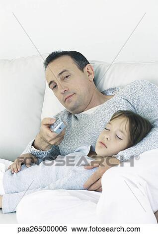 Stock Photograph - Man sitting on couch pointing remote control, holding sleeping daughter on lap. Fotosearch