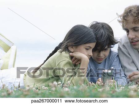 Boy and girl with father lying on stomachs on grass looking at cellphone View Large Photo Image Stock Image - Boy and girl with father lying on stomachs on grass looking at cellphone. Fotosearch