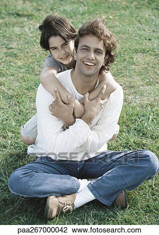 Man sitting on ground, son behind him with arms around man, both looking at camera View Large Photo Image Stock Image - Man sitting on ground, son behind him with arms around man, both looking at camera. Fotosearch