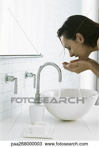 Stock Image - Woman bending over sink cupping water in hands beneath face. Fotosearch