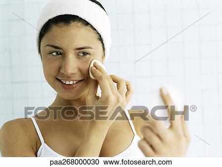 Stock Image - Woman wiping face with cotton pad, looking at compact . Fotosearch