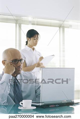 Businessman sitting at laptop with cup and saucer, woman standing behind him taking notes View Large Photo Image Picture - Businessman sitting at laptop with cup and saucer, woman standing behind him taking notes. Fotosearch