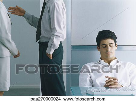 Picture - Businessman sitting back at desk with eyes closed, behind him businesspeople standing together, mid section. Fotosearch
