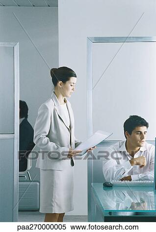 Stock Photography - Businesswoman holding document, standing next to man sitting at keyboard in office. Fotosearch