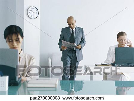 Stock Photo - In office, two women working at laptops, one man walking and reading newspaper. Fotosearch