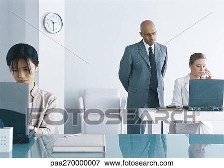Stock Photo - Two women working on laptops in office, man looking over one woman's shoulder. Fotosearch