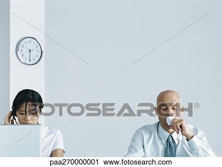 Stock Image - Woman with headset behind computer screen next to man drinking coffee. Fotosearch