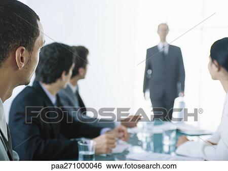 Businesspeople sitting at table with heads turned toward backlit businessman in background View Large Photo Image Stock Photograph - Businesspeople sitting at table with heads turned toward backlit businessman in background. Fotosearch