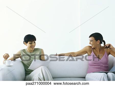 Picture - Two women sitting on sofa smiling, one with document and cup and saucer. Fotosearch