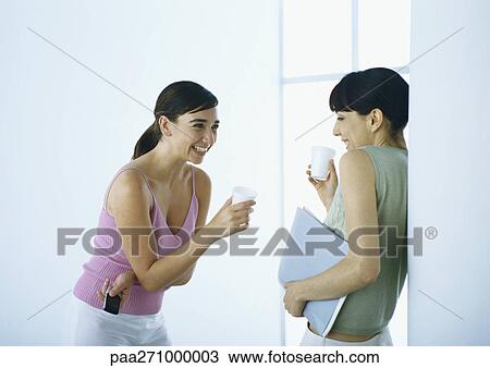 Stock Image - Two women standing with cups, laughing. Fotosearch