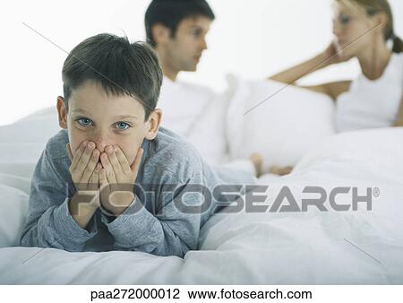 Stock Image - Boy lying on stomach in bed covering mouth with hands, parents discussing in background. Fotosearch