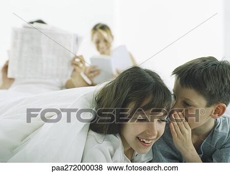 Stock Photo - Boy whispering to girl, parents reading in background. Fotosearch