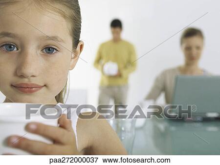 Stock Photo - Girl holding mug, parents busy in background. Fotosearch