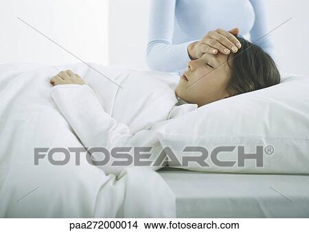 Picture - Girl lying in bed under covers, woman's hand on girl's forehead. Fotosearch