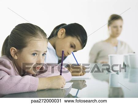 Stock Image - Two girls sitting at table writing, woman in background. Fotosearch