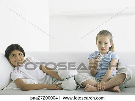 Two girls sitting on sofa, one holding remote control View Large Photo Image Stock Image - Two girls sitting on sofa, one holding remote control. Fotosearch