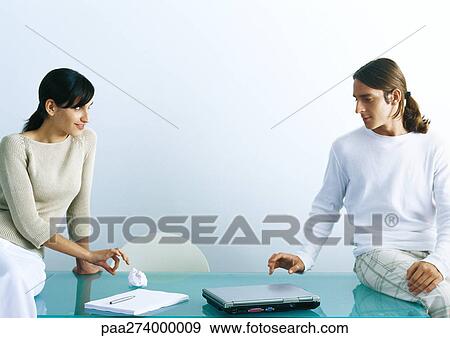 Stock Photo - Man and woman sitting on table, woman flicking paper ball to man. Fotosearch