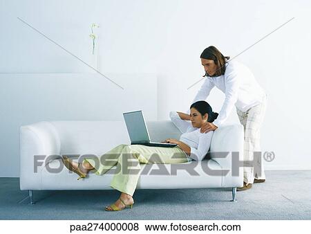 Stock Photo - Woman lounging on sofa and using laptop, man standing, looking over woman's shoulder. Fotosearch