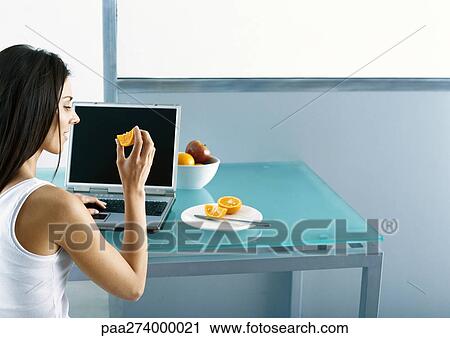 Woman sitting at table with laptop, eating fruit View Large Photo Image Stock Image - Woman sitting at table with laptop, eating fruit. Fotosearch