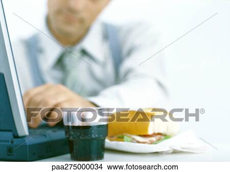 Man using laptop and having meal, focus on drink and sandwich in foreground View Large Photo Image Picture - Man using laptop and having meal, focus on drink and sandwich in foreground. Fotosearch
