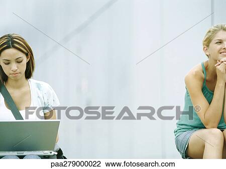 Stock Image - Two students sitting outdoors, one using laptop. Fotosearch