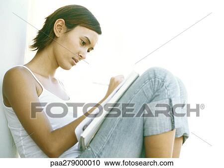 Stock Image - Young woman sitting with knees up, writing, side view. Fotosearch