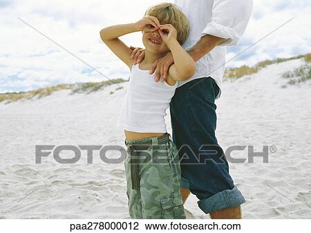 Stock Image - Boy holding hands around eyes, leaning against father on beach. Fotosearch