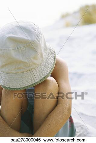 Stock Photograph - Child sitting with head down, face hidden behind hat, on beach. Fotosearch