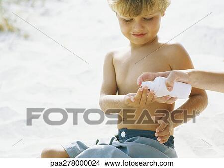 Picture - Little boy sitting on sand, woman's hand squirting sunscreen into boy's hand. Fotosearch