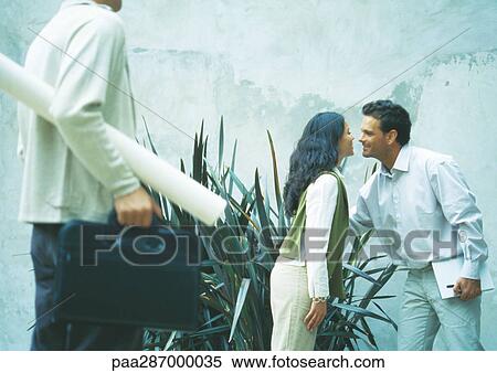 Man and woman greeting each other with cheek kiss while second man holds rolled up blueprints View Large Photo Image Stock Photography - Man and woman greeting each other with cheek kiss while second man holds rolled up blueprints. Fotosearch