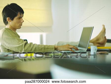 Young man sitting at table with feet up, using laptop View Large Photo Image Picture - Young man sitting at table with feet up, using laptop. Fotosearch