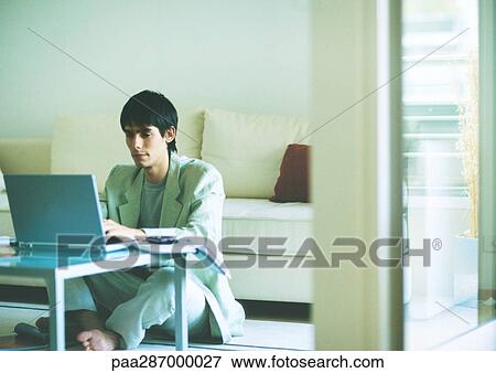 Stock Photo - Young man sitting on floor, using laptop on coffee table. Fotosearch