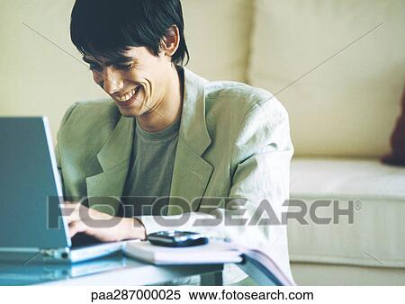 Stock Photography - Young man smiling and using laptop on coffee table. Fotosearch