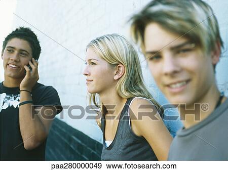Young people standing next to wall, one using cell phone View Large Photo Image Stock Photo - Young people standing next to wall, one using cell phone. Fotosearch