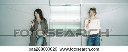 Two young women leaning against wall, looking at cell phones View Large Photo Image Stock Photograph - Two young women leaning against wall, looking at cell phones. Fotosearch